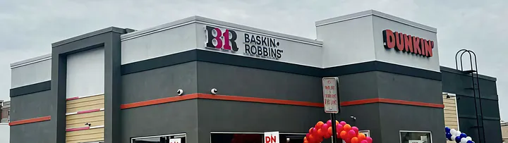 Modern co-branded corner storefront with dark gray facade and orange stripe trim, featuring BR BASKIN ROBBINS in pink and brown letters on the left tower and DUNKIN' in bold brown letters on the right tower, with red, blue, and white balloons at the entrance.