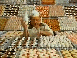 Animated GIF of Fred the Baker from vintage Dunkin' Donuts ad, wearing a white paper hat, standing in front of racks of donuts and taking a bite, with text DUNKIN DONUTS Its worth the trip.