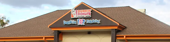 Exterior photo of a combined Dunkin' Donuts and Baskin-Robbins storefront. The building has a brown shingled roof with orange trim, and a sign on the gable shows the Dunkin' Donuts Coffee & More logo above the Baskin-Robbins BR logo.