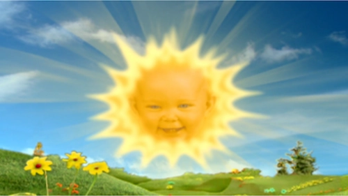 The Teletubbies baby sun with a smiling baby face in the center of a bright yellow sun shining over the green Teletubbyland landscape with yellow flowers and blue sky.