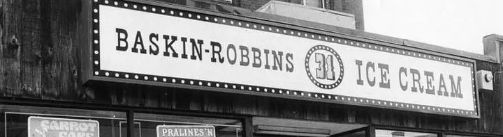 Black-and-white archival photo of a vintage Baskin-Robbins store sign. The rectangular sign reads BASKIN-ROBBINS 31 ICE CREAM in bold lettering with a light-bulb dotted border, mounted above the storefront with wood paneling. The circular 31 logo is visible in the middle.